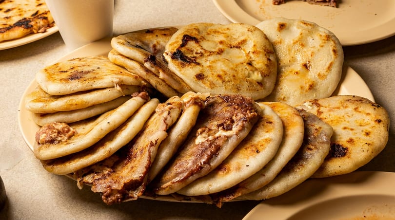 Large platter of golden-brown pupusas, some filled with meat and cheese, on a light-colored table.