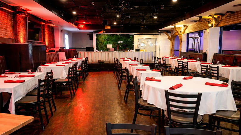 Spacious restaurant dining room with set tables, booths, and a stage, illuminated by red lights.