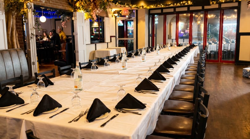 Long banquet table set with white tablecloths, black napkins, and glasses in a warm restaurant.