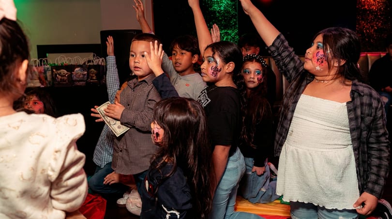 Kids with face paint raise hands at a party, one boy holding a dollar bill.
