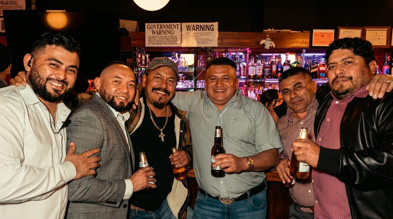 Six men smiling and holding beer bottles, embracing each other in a bar.